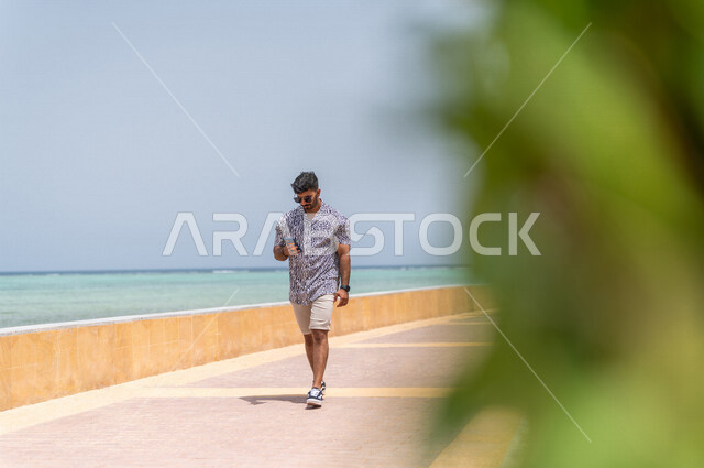 رجل عربي سعودي خليجي يمارس رياضة المشي في ممشى كورنيش جده, إستخدام الهاتف المحمول, التواصل الإجتماعي عبر الهاتف المحمول, شاطيء سياحي, التنزه في جده, الواجهه البحريه لمدينة جده