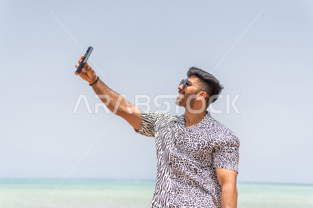 رجل عربي سعودي خليجي يقوم بإلتقاط صور تذكاريه في ممشى كورنيش جده, تصوير سلفي, إستخدام الهاتف المحمول, شاطيء سياحي, التنزه في جده, الواجهه البحريه لمدينة جده