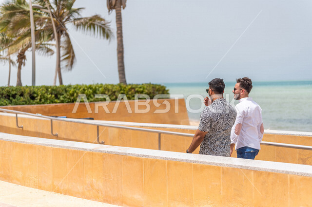 Two Saudi Arabian Gulf friends walking on the promenade of the Jeddah Corniche, a tourist beach, outdoor physical activity, hiking in Jeddah, chatting and conversations, having fun with friends, the waterfront of Jeddah