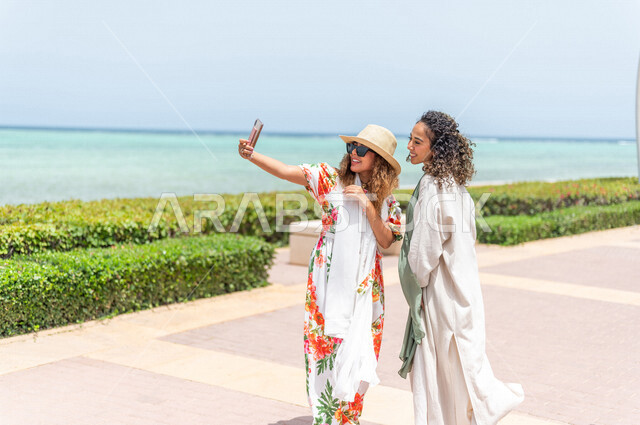 صديقتان عربيتان سعوديتان خليجيتان تلتقطان صور تذكاريه في ممشى كورنيش جده, إستخدام الهاتف المحمول, شاطيء سياحي, التنزه في جده, دردشة ومحادثات, الإستمتاع مع الأصدقاء, الواجهه البحريه لمدينة جده, عطلة صيفيه
