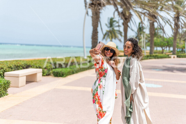 صديقتان عربيتان سعوديتان خليجيتان تلتقطان صور تذكاريه في ممشى كورنيش جده, إستخدام الهاتف المحمول, شاطيء سياحي, التنزه في جده, دردشة ومحادثات, الإستمتاع مع الأصدقاء, الواجهه البحريه لمدينة جده, عطلة صيفيه