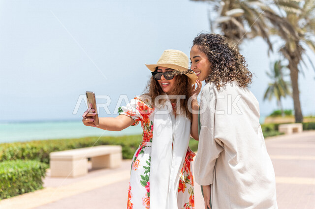 صديقتان عربيتان سعوديتان خليجيتان تلتقطان صور تذكاريه في ممشى كورنيش جده, إستخدام الهاتف المحمول, شاطيء سياحي, التنزه في جده, دردشة ومحادثات, الإستمتاع مع الأصدقاء, الواجهه البحريه لمدينة جده, عطلة صيفيه