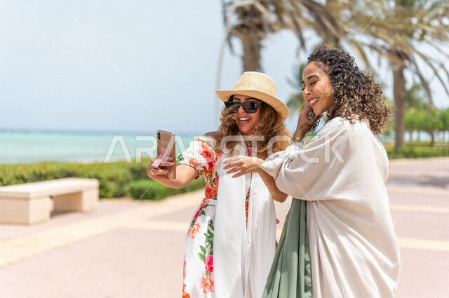 صديقتان عربيتان سعوديتان خليجيتان تلتقطان صور تذكاريه في ممشى كورنيش جده, إستخدام الهاتف المحمول, شاطيء سياحي, التنزه في جده, دردشة ومحادثات, الإستمتاع مع الأصدقاء, الواجهه البحريه لمدينة جده, عطلة صيفيه