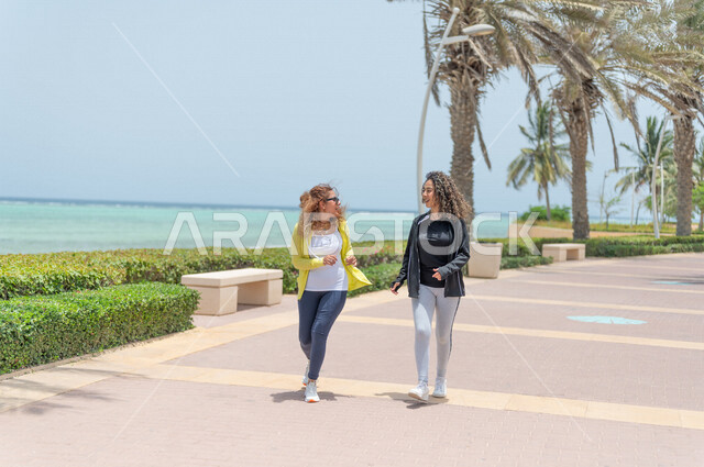 صديقتان عربيتان سعوديتان خليجيتان تمارسان رياضة المشي في ممشى كورنيش جده, شاطيء سياحي, نشاط بدني في الهواء الطلق, التنزه في جده, دردشة ومحادثات, الإستمتاع مع الأصدقاء, الواجهه البحريه لمدينة جده
