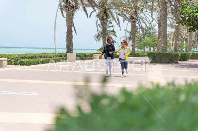 صديقتان عربيتان سعوديتان خليجيتان تمارسان رياضة المشي في ممشى كورنيش جده, شاطيء سياحي, نشاط بدني في الهواء الطلق, التنزه في جده, دردشة ومحادثات, الإستمتاع مع الأصدقاء, الواجهه البحريه لمدينة جده