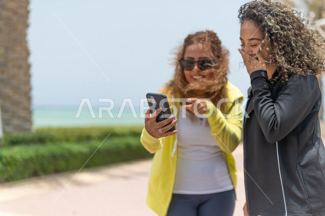 صديقتان عربيتان سعوديتان خليجيتان في ممشى كورنيش جده, إستخدام الهاتف المحمول, التواصل الإجتماعي, شاطيء سياحي, التنزه في جده, دردشة ومحادثات, الإستمتاع مع الأصدقاء, الواجهه البحريه لمدينة جده