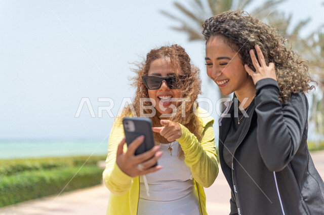 صديقتان عربيتان سعوديتان خليجيتان في ممشى كورنيش جده, إستخدام الهاتف المحمول, التواصل الإجتماعي, شاطيء سياحي, التنزه في جده, دردشة ومحادثات, الإستمتاع مع الأصدقاء, الواجهه البحريه لمدينة جده