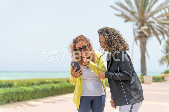 صديقتان عربيتان سعوديتان خليجيتان في ممشى كورنيش جده, إستخدام الهاتف المحمول, التواصل الإجتماعي, شاطيء سياحي, التنزه في جده, دردشة ومحادثات, الإستمتاع مع الأصدقاء, الواجهه البحريه لمدينة جده
