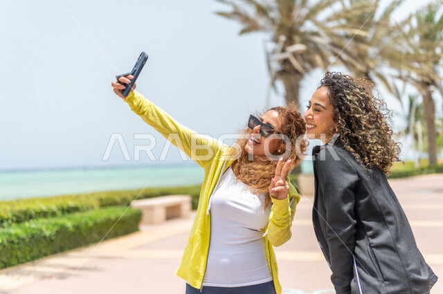 صديقتان عربيتان سعوديتان خليجيتان تلتقطان صور تذكاريه في ممشى كورنيش جده, إستخدام الهاتف المحمول, شاطيء سياحي, التنزه في جده, دردشة ومحادثات, الإستمتاع مع الأصدقاء, الواجهه البحريه لمدينة جده