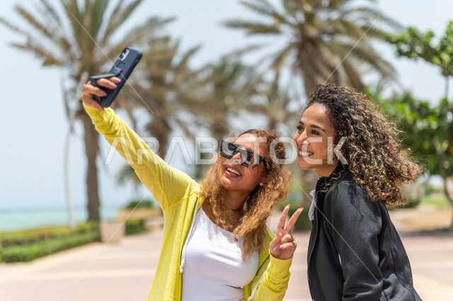 صديقتان عربيتان سعوديتان خليجيتان تلتقطان صور تذكاريه في ممشى كورنيش جده, إستخدام الهاتف المحمول, شاطيء سياحي, التنزه في جده, دردشة ومحادثات, الإستمتاع مع الأصدقاء, الواجهه البحريه لمدينة جده