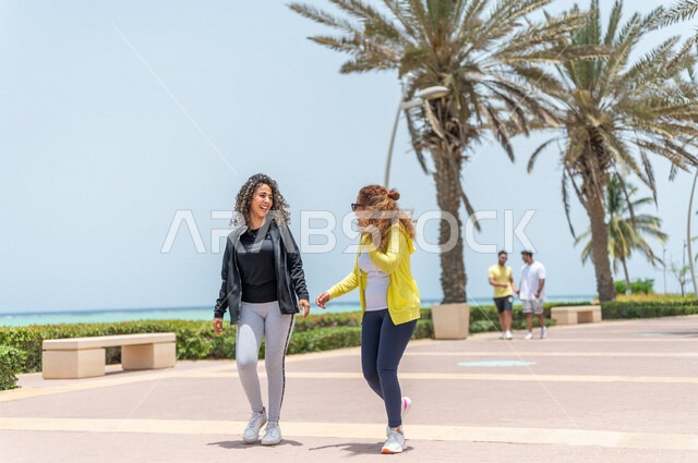 Two Saudi Arabian Gulf friends practicing walking on the Jeddah Corniche promenade, a tourist beach, outdoor physical activity, hiking in Jeddah, chatting and conversations, having fun with friends, the waterfront of Jeddah