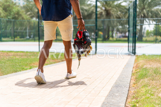 زاوية من الخلف لرجل عربي خليجي سعودي، يحمل بيده الزلاجات الدوارة في إحدى الحدائق الترفيهية بالسعودية، أماكن سياحية ترفيهية، قضاء أوقات ممتعة، أنشطة ترفيهية، ممارسة رياضة تزلج بالعجلات