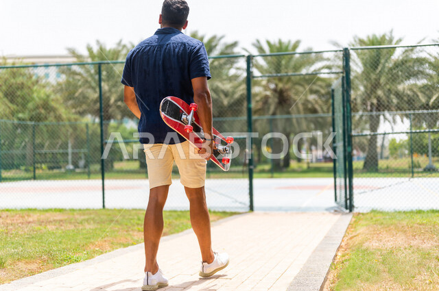 زاوية من الخلف لرجل عربي خليجي سعودي، يحمل بيده لوح التزلج في إحدى الحدائق الترفيهية بالسعودية، أماكن سياحية ترفيهية، قضاء أوقات ممتعة، أنشطة ترفيهية، ممارسة رياضة التزلج على اللوح