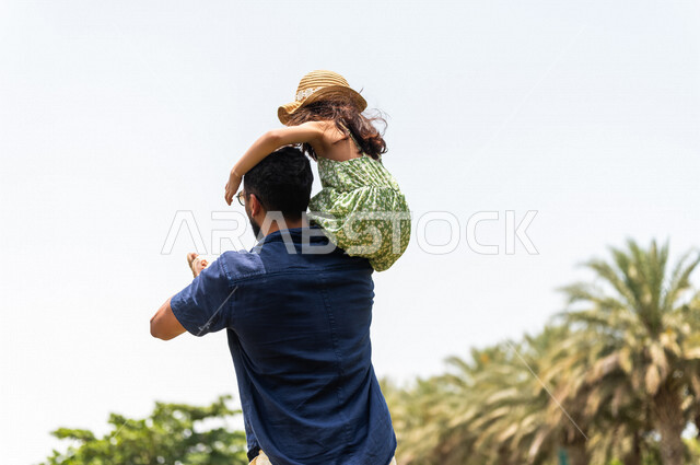 زاوية من الخلف لرجل عربي خليجي سعودي يقضي وقت ممتع مع إبنته في إحدى الحدائق الترفيهية بالسعودية، فعاليات ترفيهية عائلية، عطلة صيفية، قضاء أوقات عائلية ممتعة، الأب يحمل إبنته إلى الأعلى، حدائق ونزهات ترفيهية للعائلة في المملكة العربية السعودية