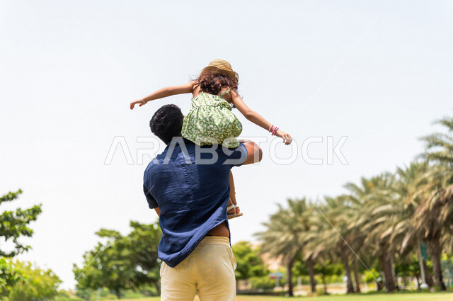 زاوية من الخلف لرجل عربي خليجي سعودي يقضي وقت ممتع مع إبنته في إحدى الحدائق الترفيهية بالسعودية، فعاليات ترفيهية عائلية، عطلة صيفية، حدائق ونزهات ترفيهية للعائلة في المملكة العربية السعودية، قضاء أوقات عائلية ممتعة، الأب يحمل إبنته إلى الأعلى