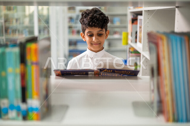 طالب عربي سعودي خليجي يقوم بقراءة كتاب من المكتبة المدرسية، الاستمتاع بقراءة الكتب، مفهوم القراءه والثقافه, مكتبة المدرسة، التعليم الذاتي, بيئه مدرسيه تعليميه متطوره, مدارس أكاديميه في المملكة العربية السعودية