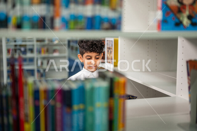 طالب عربي سعودي خليجي يقوم بإختيار كتاب من المكتبة المدرسية، الاستمتاع بقراءة الكتب، مفهوم القراءه والثقافه, مكتبة المدرسة، التعليم الذاتي, بيئه مدرسيه تعليميه متطوره, مدارس أكاديميه في المملكة العربية السعودية