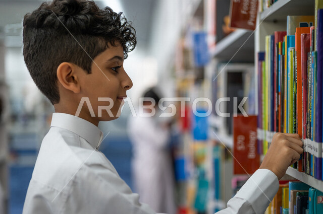 صوره مقربه لطالب عربي سعودي خليجي يقوم بإختيار كتاب من المكتبة المدرسية، الاستمتاع بقراءة الكتب، مفهوم القراءه والثقافه, مكتبة المدرسة، التعليم الذاتي, بيئه مدرسيه تعليميه متطوره, مدارس أكاديميه في المملكة العربية السعودية