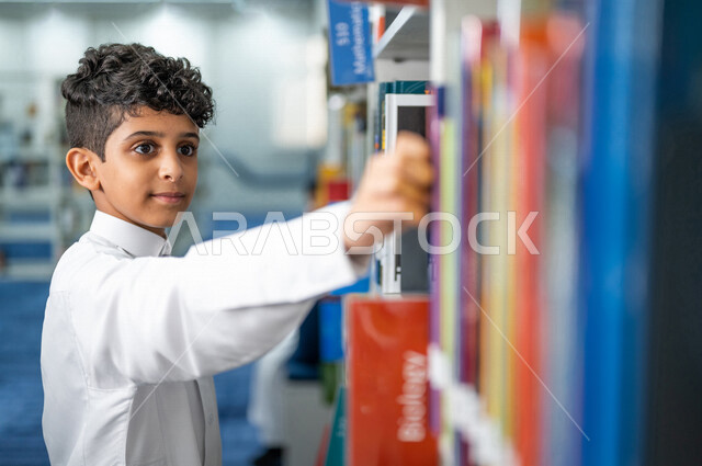 صوره مقربه لطالب عربي سعودي خليجي يقوم بإختيار كتاب من المكتبة المدرسية، الاستمتاع بقراءة الكتب، مفهوم القراءه والثقافه, مكتبة المدرسة، التعليم الذاتي, بيئه مدرسيه تعليميه متطوره, مدارس أكاديميه في المملكة العربية السعودية