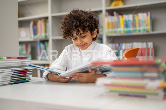 طالب عربي سعودي خليجي يقوم بقراءة كتاب من المكتبة المدرسية، الاستمتاع بقراءة الكتب، مفهوم القراءه والثقافه, مكتبة المدرسة، التعليم الذاتي, بيئه مدرسيه تعليميه متطوره, مدارس أكاديميه في المملكة العربية السعودية