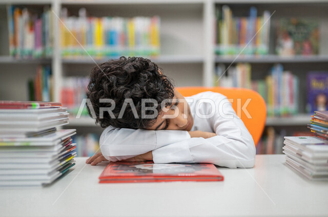 طالب عربي سعودي خليجي في حالة نوم بعد مجهود القراءه في المكتبة المدرسية، النوم والنعاس, الحاجه إلى الراحه, إيماءات النعاس والتعب, الشعور بالتعب, الحاجه الى القيلوله, مكتبة المدرسة