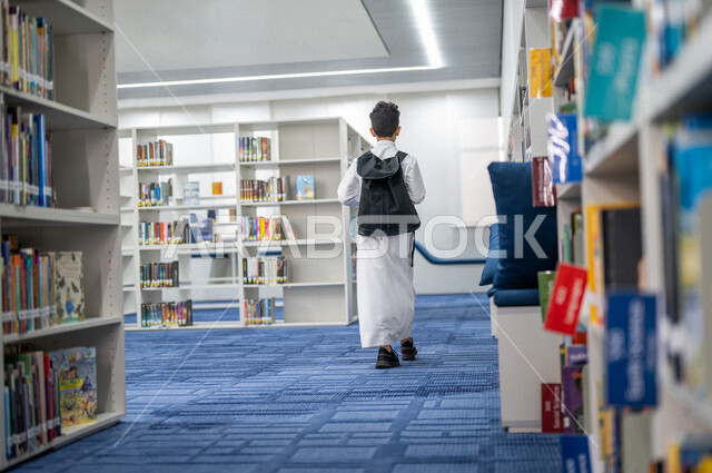 تصوير من الخلف لطالب عربي سعودي خليجي يمشي في المكتبة المدرسية، الاستمتاع بقراءة الكتب المدرسية، مفهوم التعليم والثقافه, مكتبة المدرسة، التعليم الذاتي, بيئه مدرسيه تعليميه متطوره, مدارس أكاديميه في المملكة العربية السعودية