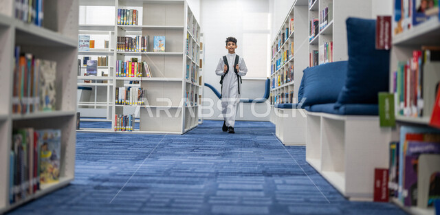 طالب عربي سعودي خليجي يمشي في المكتبة المدرسية، الاستمتاع بقراءة الكتب المدرسية، مفهوم التعليم والثقافه, مكتبة المدرسة، التعليم الذاتي, بيئه مدرسيه تعليميه متطوره, مدارس أكاديميه في المملكة العربية السعودية