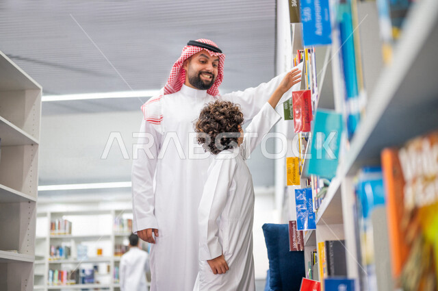 طالب عربي سعودي خليجي يقوم بإختيار كتاب من المكتبة المدرسية بمساعدة المعلم، الاستمتاع بقراءة الكتب، مفهوم القراءه والثقافه, مكتبة المدرسة، التعليم الذاتي, بيئه مدرسيه تعليميه متطوره, مدارس أكاديميه في المملكة العربية السعودية