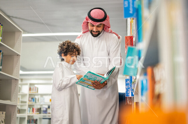 طالب عربي سعودي خليجي يقوم بقراءة كتاب بمساعدة المعلم في المكتبة المدرسية، الاستمتاع بقراءة الكتب، مفهوم القراءه والثقافه, مكتبة المدرسة، التعليم الذاتي, بيئه مدرسيه تعليميه متطوره, مدارس أكاديميه في المملكة العربية السعودية