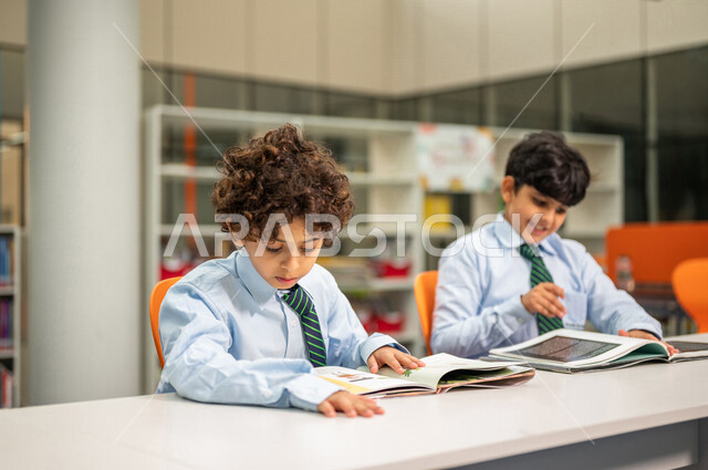 طالبان عربيان سعوديان خليجيان يجلسان في المكتبة المدرسية، الاستمتاع بقراءة الكتب، مفهوم التعليم الذاتي, مكتبة المدرسة، بيئه مدرسيه تعليميه متطوره, المدارس المبتكره, جودة التعليم, مدارس أكاديميه في المملكة العربية السعودية