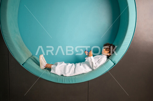 طالب عربي سعودي خليجي يتمدد على مقعد مريح, يتناول البسكويت في وقت الإستراحه, وجبه خفيفه, وقت الفسحه المدرسيه, بيئه تعليميه مدرسيه حديثه متطوره, مدارس أكاديميه في المملكه العربيه السعوديه