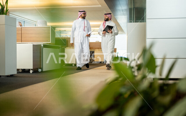 موظفان عربيان سعوديان خليجيان داخل مقر الشركة، يرتديان الزي السعودي التقليدي، مهن و وظائف سعودية، شركة سعودية، رجال أعمال، الحوارات والنقاشات، بيئة العمل، دردشة عمل، متابعة ومناقشة الأعمال، زملاء عمل، مناقشة ومتابعة الأهداف،