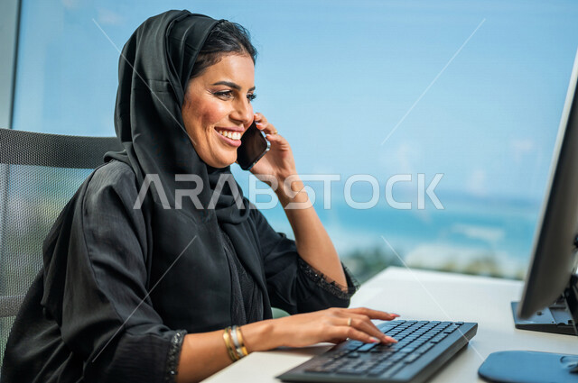 مرأة عربية خليجية سعودية، تقوم بإجراء مكالمة هاتفية عبر الجوال، إستخدام جهاز الحاسوب المحمول، مهن ووظائف سعودية، متابعة ومناقشة الأعمال، عقد إجتماع عمل عبر الإنترنت، التسويق الإلكتروني، تصفح الإنترنت، شركة سعودية، العمل عن بعد