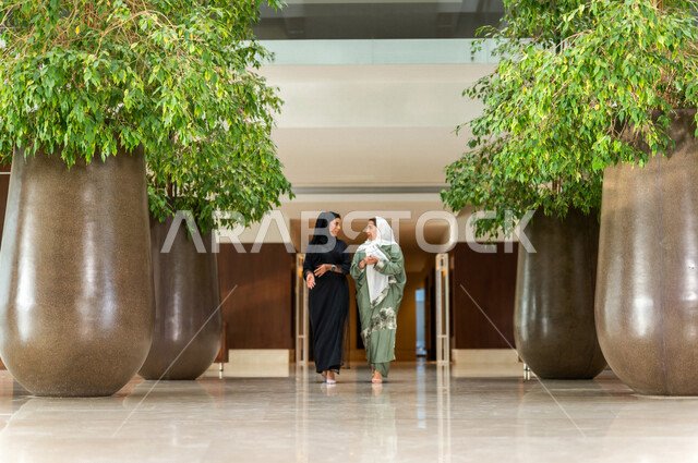 Two Saudi Arabian Gulf employees inside the company's headquarters, wearing distinctive modern abayas, veiled fashion, Saudi professions and jobs, a Saudi company, business women, dialogues and discussions, work environment, work chat