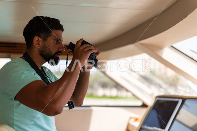 صوره مقربه لرجل عربي سعودي خليجي في قمرة القياده في يخت وسط البحر, إستخدام المنظار لتقريب الرؤيه, مشاهدة المناظر الطبيعيه عبر المنظور, قيادة اليخوت, متعة الإبحار في القوارب والسفن في البحر , جولات بحريه ممتعه, أنشطه ترفيهيه, رحلات بحريه, عطله صيفيه