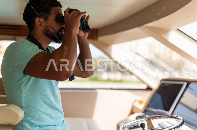 صوره مقربه لرجل عربي سعودي خليجي في قمرة القياده في يخت وسط البحر, إستخدام المنظار لتقريب الرؤيه, مشاهدة المناظر الطبيعيه عبر المنظور, قيادة اليخوت, متعة الإبحار في القوارب والسفن في البحر , جولات بحريه ممتعه, أنشطه ترفيهيه, رحلات بحريه, عطله صيفيه