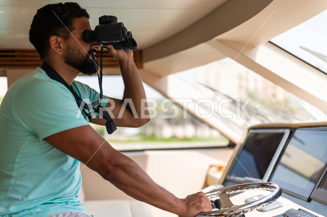 صوره مقربه لرجل عربي سعودي خليجي في قمرة القياده في يخت, التحكم بعجلة القياده, توجيه المقود, إستخدام المنظار لتقريب الرؤيه, مشاهدة المناظر الطبيعيه عبر المنظور, قيادة اليخوت, متعة الإبحار في القوارب والسفن, رحلات و جولات بحريه, أنشطه ترفيهيه, عطله صيفيه