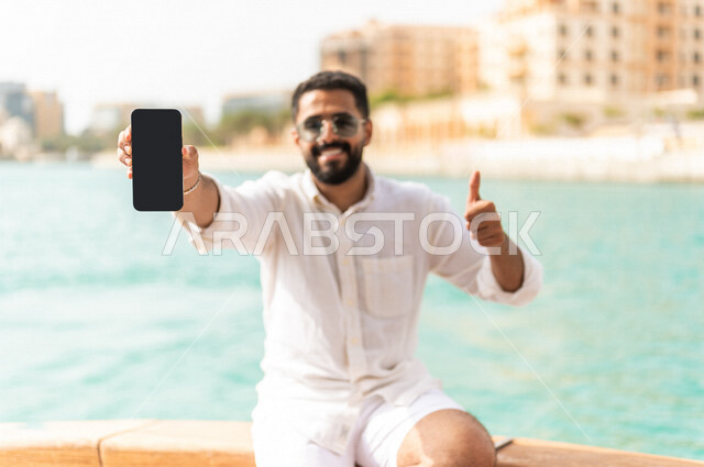 شاب عربي سعودي خليجي على متن يخت في البحر, يعرض شاشة سوداء فارغه على الهاتف المحمول, التسويق الرقمي, تطبيقات الهاتف المحمول, متعة الإبحار في القوارب والسفن, جولات بحريه على كورنيش جده, رحلات بحريه في مارينا جده, عطله صيفيه, السياحه في السعوديه