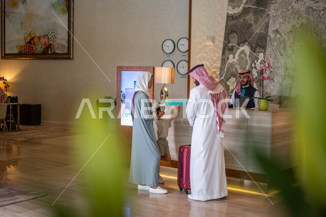 صورة من الخلف لعائلة عربية خليجية سعودية تقف امام مكتب موظفين الإستقبال في فندق سياحي، اتمام إجراءات الحجز والدخول لدى مكتب استقبال العملاء في الفندق، خدمة العملاء، حقيبة سفر حمراء، سياحة وفندقة، رحلة عائلية سعيدة، فنادق فاخرة في السعودية