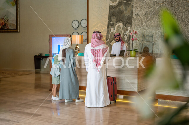 صورة من الخلف لعائلة عربية خليجية سعودية تقف امام مكتب موظفين الإستقبال في فندق سياحي، اتمام إجراءات الحجز والدخول لدى مكتب استقبال العملاء في الفندق، خدمة العملاء، حقيبة سفر حمراء، سياحة وفندقة، رحلة عائلية سعيدة، فنادق فاخرة في السعودية