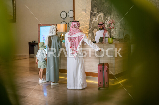 صورة من الخلف لعائلة عربية خليجية سعودية تقف امام مكتب موظفين الإستقبال في فندق سياحي، اتمام إجراءات الحجز والدخول لدى مكتب استقبال العملاء في الفندق، خدمة العملاء، حقيبة سفر حمراء، سياحة وفندقة، رحلة عائلية سعيدة، فنادق فاخرة في السعودية