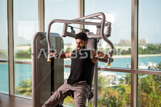 رجل عربي خليجي سعودي يرتدي الملابس الرياضية ويقوم بالتمارين الرياضية، استخدام الأجهزة، زي رياضي، تقوية وبناء العضلات، ممارسة تمارين رفع الاوزان، صالة الألعاب الرياضية، انشطة وتمارين رياضية، الصحة واللياقة البدنية، نشاط بدني