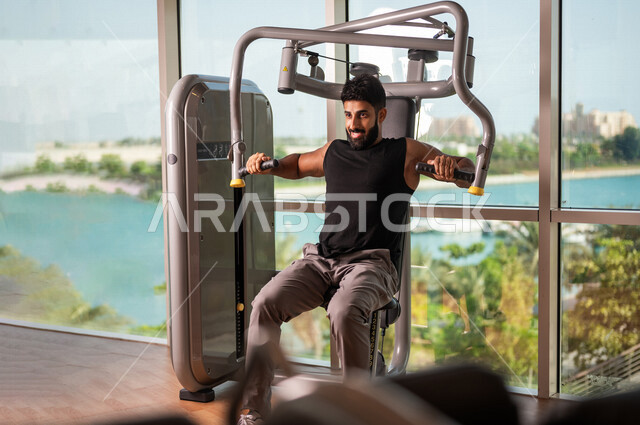 رجل عربي خليجي سعودي يرتدي الملابس الرياضية ويقوم بالتمارين الرياضية، استخدام الأجهزة، زي رياضي، تقوية وبناء العضلات، ممارسة تمارين رفع الاوزان، صالة الألعاب الرياضية، انشطة وتمارين رياضية، الصحة واللياقة البدنية، نشاط بدني
