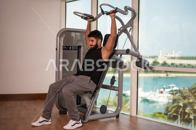 رجل عربي خليجي سعودي يرتدي الملابس الرياضية ويقوم بالتمارين الرياضية، استخدام الأجهزة، زي رياضي، تقوية وبناء العضلات، ممارسة تمارين رفع الاوزان، صالة الألعاب الرياضية، انشطة وتمارين رياضية، الصحة واللياقة البدنية، نشاط بدني