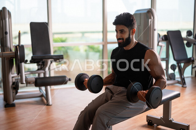 رجل عربي خليجي سعودي يرتدي الملابس الرياضية ويقوم بالتمارين الرياضية، استخدام الدمبل، زي رياضي، تقوية وبناء العضلات، ممارسة تمارين رفع الاوزان، صالة الألعاب الرياضية، انشطة وتمارين رياضية، الصحة واللياقة البدنية، نشاط بدني