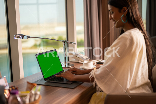 A Saudi Arabian Gulf woman using a laptop with a green chroma screen, making a video call, completing work and tasks, live video broadcasting, chatting and communication, green chroma, working remotely, summer vacation, dating and socializing, online frie