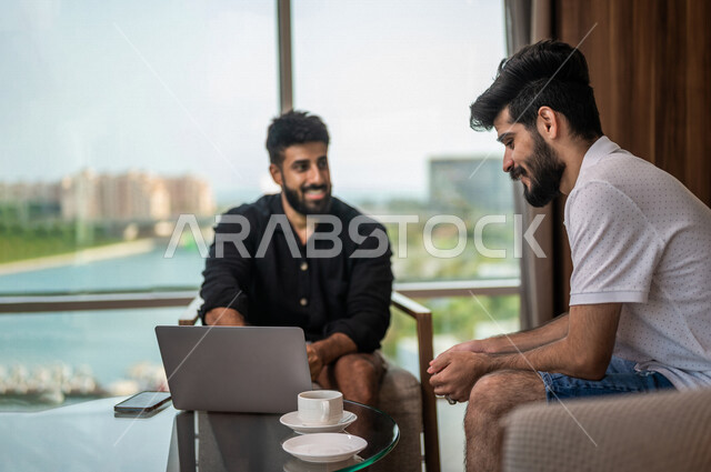 Two smiling Saudi Arabian Gulf men sitting in the cafe, enjoying hot coffee, using a laptop, dialogue and discussion, a cup of coffee, hot drinks, Saudi youth office, friendship session, youth atmosphere in a Saudi cafe