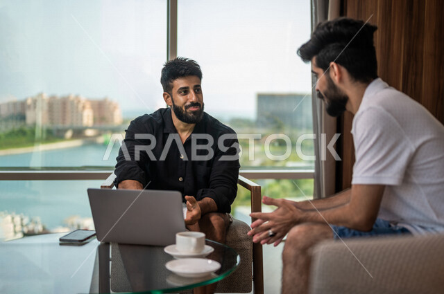 شابان عربيان خليجيان سعوديان مبتسمان يجلسان في المقهى، الاستمتاع بشرب القهوة الساخنة، استخدام جهاز الحاسوب المحمول، حوار ونقاش، كوب قهوة، مشروبات ساخنة، ديوانية شباب السعودية، جلسة صداقة، أجواء شبابية في مقهى سعودي