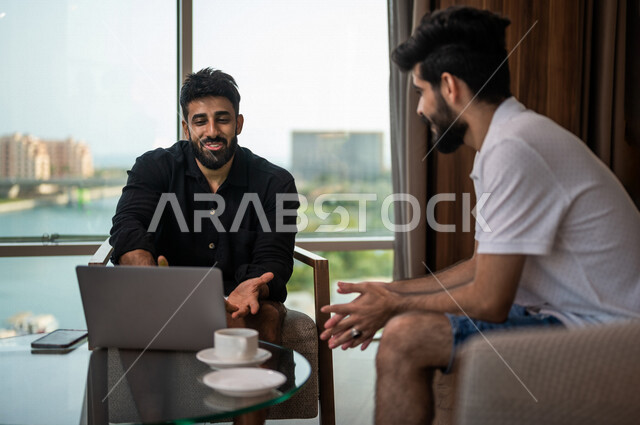 شابان عربيان خليجيان سعوديان مبتسمان يجلسان في المقهى، الاستمتاع بشرب القهوة الساخنة، استخدام جهاز الحاسوب المحمول، حوار ونقاش، كوب قهوة، مشروبات ساخنة، ديوانية شباب السعودية، جلسة صداقة، أجواء شبابية في مقهى سعودي