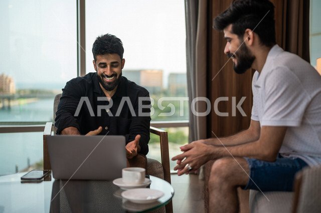 شابان عربيان خليجيان سعوديان مبتسمان يجلسان في المقهى، الاستمتاع بشرب القهوة الساخنة، استخدام جهاز الحاسوب المحمول، حوار ونقاش، كوب قهوة، مشروبات ساخنة، ديوانية شباب السعودية، جلسة صداقة، أجواء شبابية في مقهى سعودي
